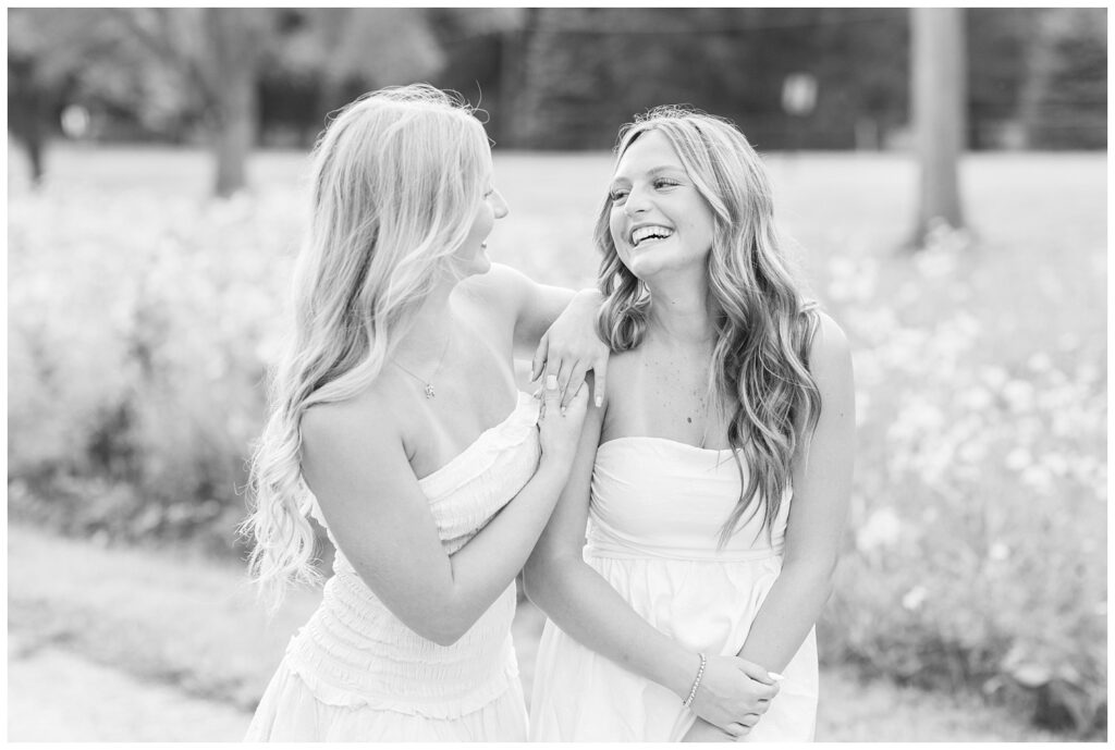 two senior best friends wearing white dresses and laughing with each other next to wildflowers