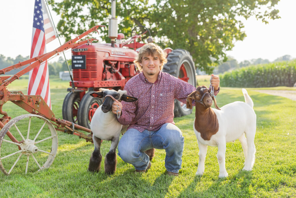 high school senior squatting in front of his tractor outside holding goats