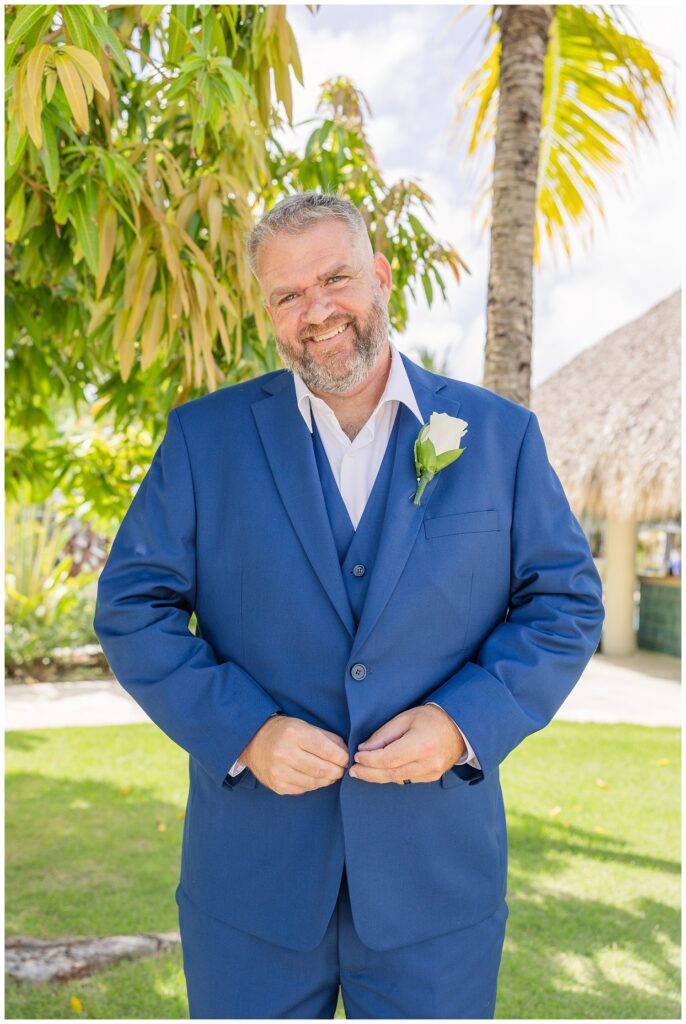 groom smiling while adjusting his jacket buttons while wearing a blue suit and white rose boutonniere 