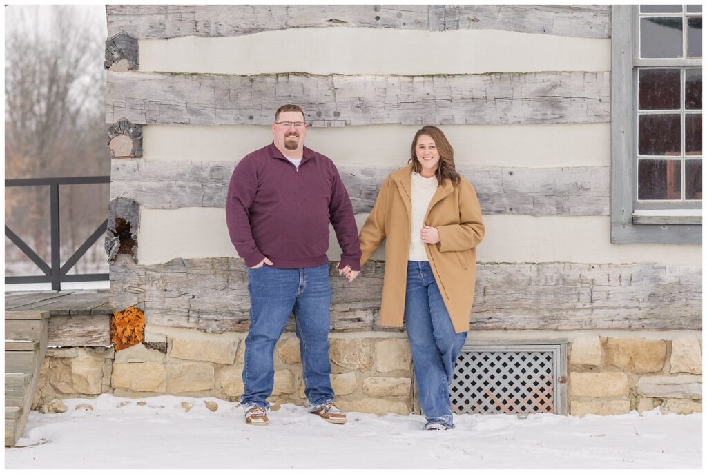 engaged couple holding hands and leaning against a log cabin in the snow