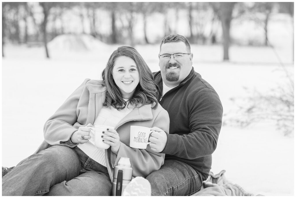 engaged couple sitting together on a blanket holding coffee mugs