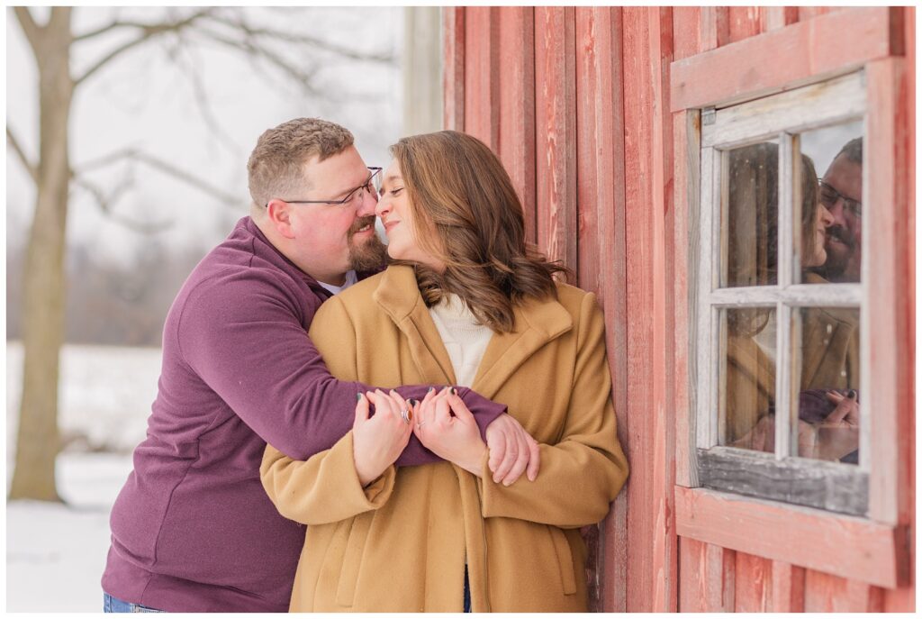 fiance hugging his fiancee from behind and touching noses against a red barn in the snow