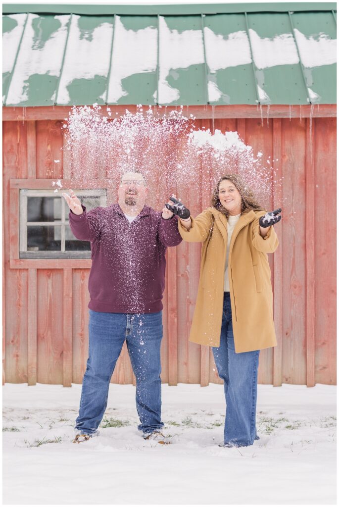 engaged couple throwing up balls of snow standing in front of a red barn in Bloomville, Ohio