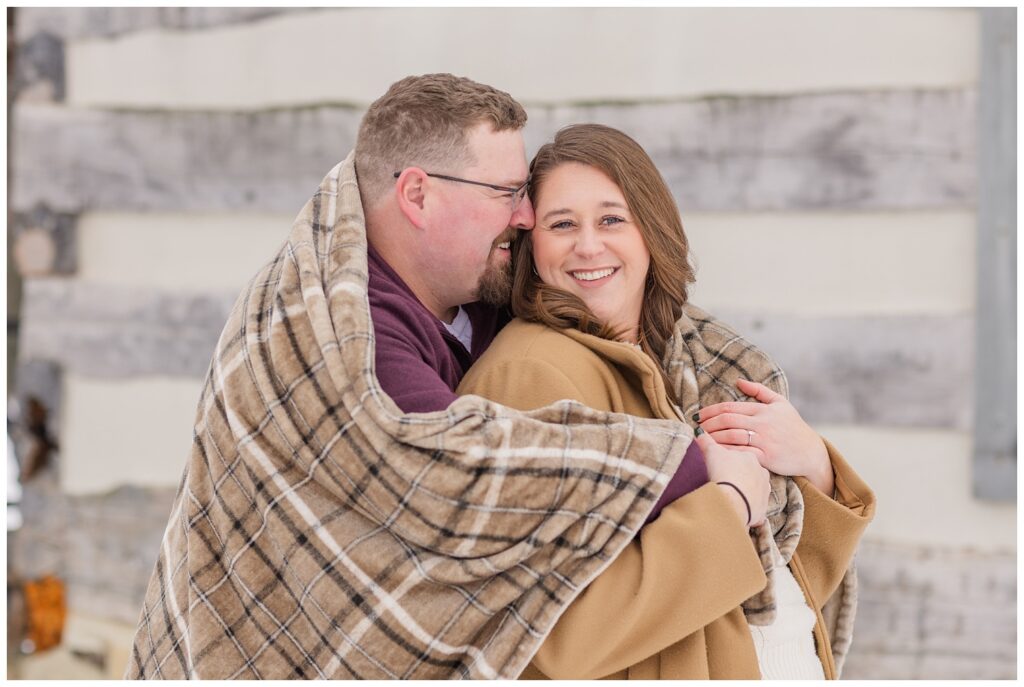 couple snuggling in a blanket in front of a log cabin with Bloomville, OH engagement photographer