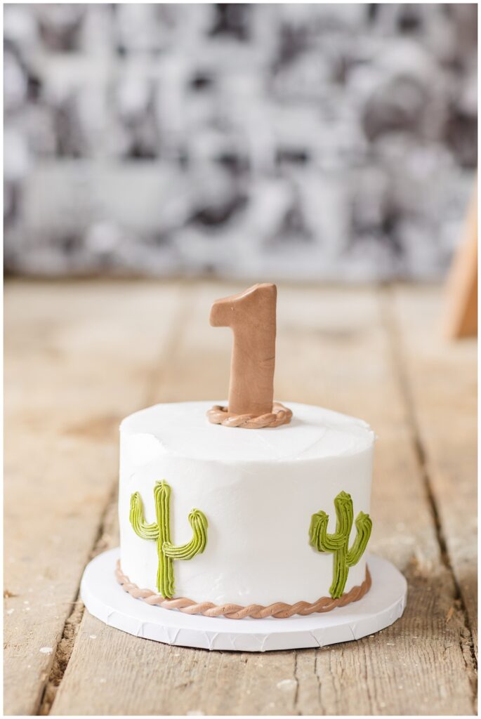 white smash cake decorated with green cacti and a number one on top on a wood floor
