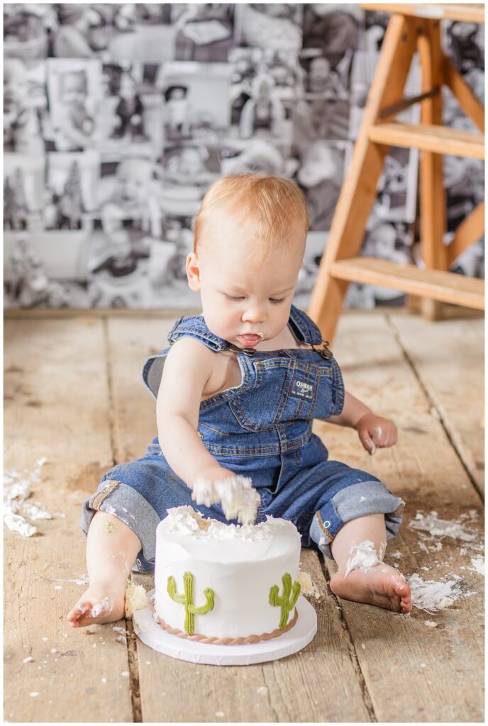 one year old boy digging into his white cacti decorated smash cake on a wooden floor 