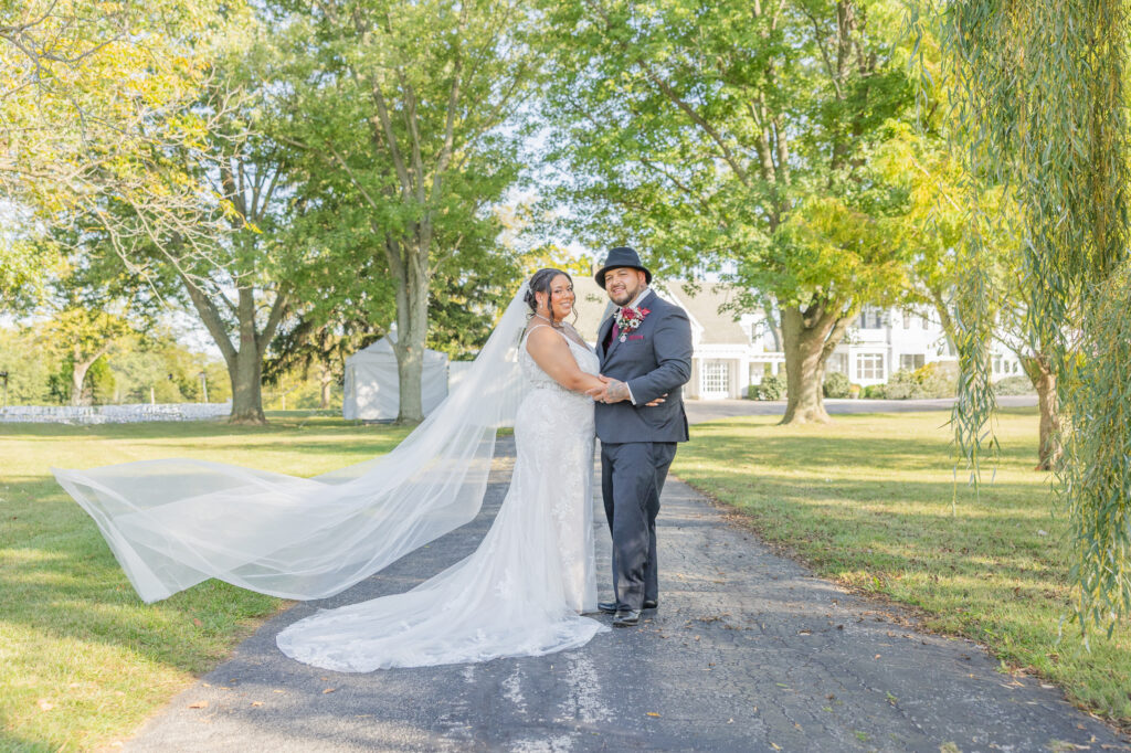 wedding couple posing on a driveway while the bride's veil flies in the open air