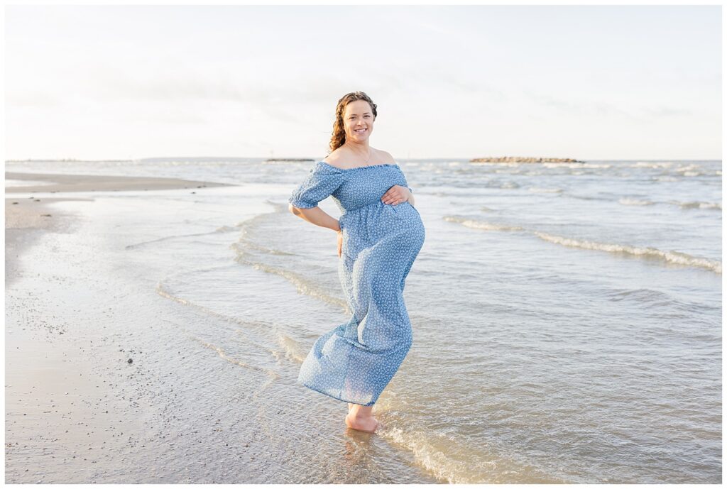 mom to be holding her hand over the top of her pregnant belly wearing a blue dress while standing in the ocean