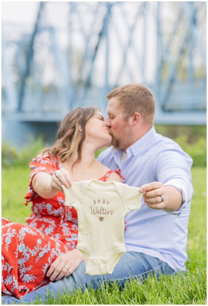 married couple showing new baby onesie and kissing at Tindall Bridge in Ohio