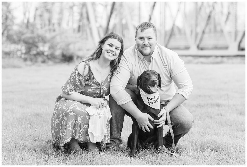 married couple showing their baby onesie and their dog with a big brother bandana at pregnancy portrait session